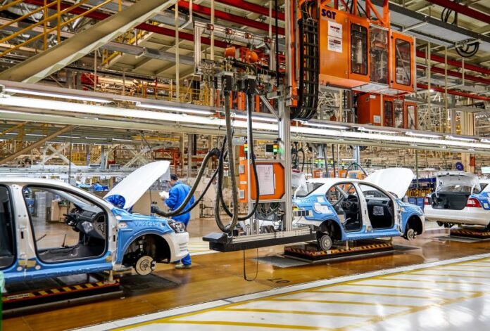 White and blue car frames moving on an assembly line in an expansive factory hangar.