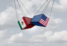 Two shipping containers, one with a U.S. flag and one with a Mexican flag, suspended next to each other in the air by thick cables. They are crashing into each other in mid air.