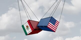 Two shipping containers, one with a U.S. flag and one with a Mexican flag, suspended next to each other in the air by thick cables. They are crashing into each other in mid air.