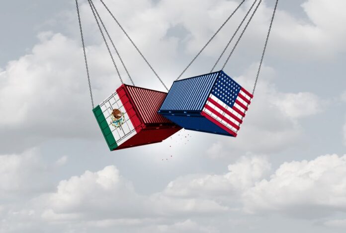 Two shipping containers, one with a U.S. flag and one with a Mexican flag, suspended next to each other in the air by thick cables. They are crashing into each other in mid air.