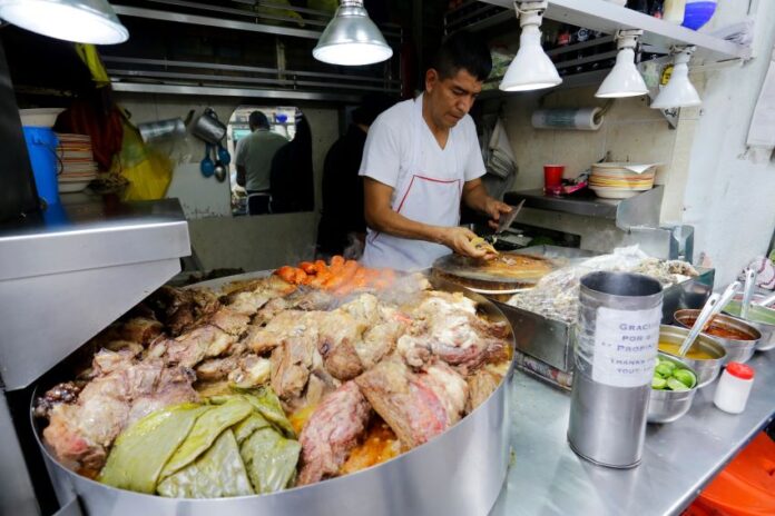 A Mexico City taquero prepares food in the background, with a pile of roasted nopales, meat and vegetables in the foreground