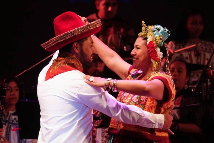 A man and a woman in traditional Oaxacan costumes dancing together. The woman is wearing a tiara of flowers while the man wears a traditional red Oaxacan hat, a smaller variation of the Mexican sombrero. They are smiling at each other as they dance.