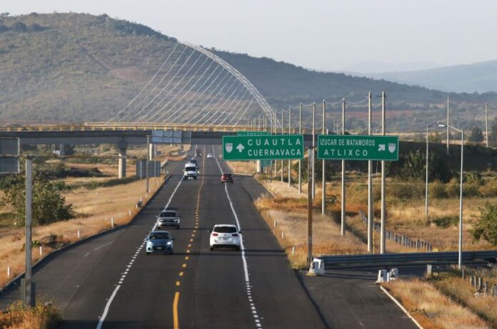 A federal divided highway in Mexico with one lane in each direction. Cars are traveling in both directions near a highway signs for Cuautla and Cuernavaca and an exit sign for Izucar de Matamoros and Atlixco.