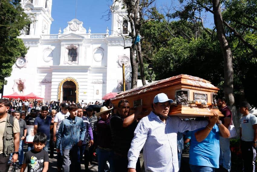 Funeral procession for man in Chilpancingo, Guerrero, who was a victim of homicide in the downtown of the city. Mexican men are at the front of the photo, acting as pallbearers and carrying a wooden coffin on their shoulders through the street from a church. Behind them walk family and friends in mourning.