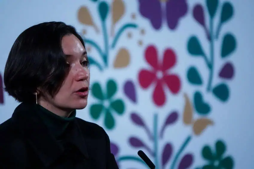 A woman with black hair and gold earrings standing in side profile at President Sheinbaum's press conference.