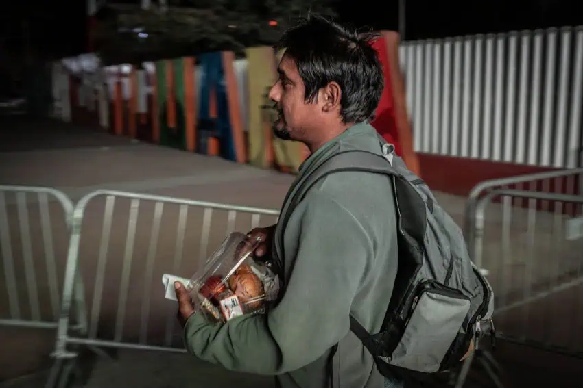 A young man in a gray hoodie-style sweatshirt and wearing a backpack carries a clamshell plastic food container in his arms as he passes temporary gates at the Mexico-US border into Tijuana. Off to the side in the background of the photo is a larger than life letter sculpture of the word "Tijuana"