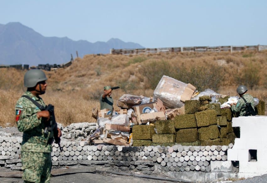 Mexican army soldiers standing near a pile of boxes and bales of marijuana sitting on top of each other atop rows of dried wood to create a bonfire to burn the drugs. They are standing in an open outdoor area in Nuevo Leon, Mexico, filled with dried grass and scrub plants.