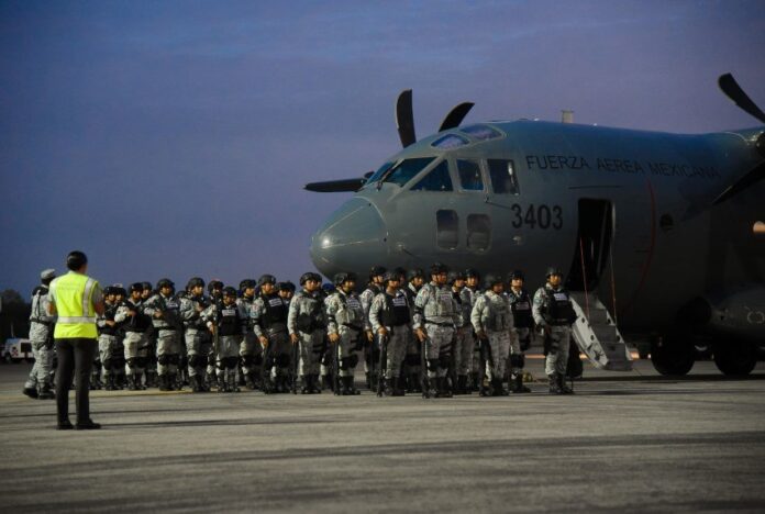 National Guardsmen in gray uniforms at dawn, standing in formation next to a Mexican Airforce carrier jet, waiting to board.