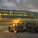 Members of Mexico's National Guard waiting with their belongings outside a troop carrier plane that has the name Guardia Nacional and the logo of Mexico's armed forces on it. The top half of the plane is painted olive drab and the bottom half of the plane is cream colored. It is early morning, just after dawn.