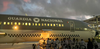 Members of Mexico's National Guard waiting with their belongings outside a troop carrier plane that has the name Guardia Nacional and the logo of Mexico's armed forces on it. The top half of the plane is painted olive drab and the bottom half of the plane is cream colored. It is early morning, just after dawn.