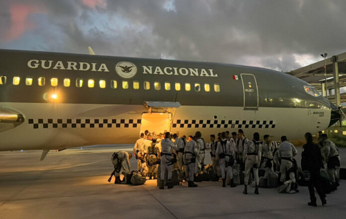 Members of Mexico's National Guard waiting with their belongings outside a troop carrier plane that has the name Guardia Nacional and the logo of Mexico's armed forces on it. The top half of the plane is painted olive drab and the bottom half of the plane is cream colored. It is early morning, just after dawn.