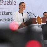 President Claudia Sheinbaum smiling as she stands behind the presidential podium at her daily press conference. She's wearing a white blazer and a black under blouse and looking out at reporters.