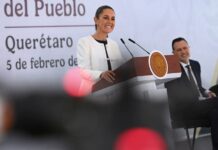 President Claudia Sheinbaum smiling as she stands behind the presidential podium at her daily press conference. She's wearing a white blazer and a black under blouse and looking out at reporters.