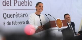 President Claudia Sheinbaum smiling as she stands behind the presidential podium at her daily press conference. She's wearing a white blazer and a black under blouse and looking out at reporters.