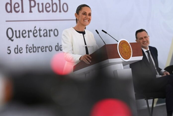 President Claudia Sheinbaum smiling as she stands behind the presidential podium at her daily press conference. She's wearing a white blazer and a black under blouse and looking out at reporters.