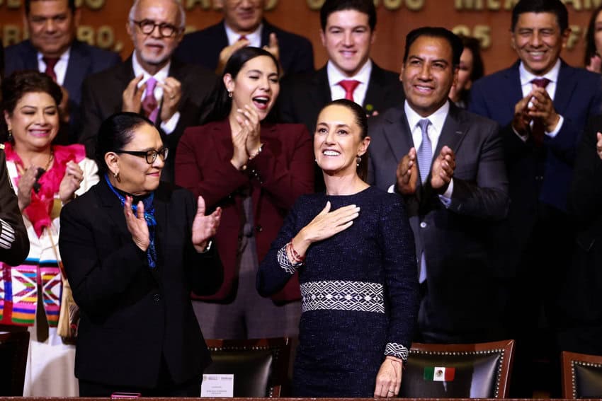 Mexico's President Claudia Sheinbaum in a dark colored dress with a black-and-white traditional Mexican Indigenous pattern around the waistline and cuffs. She is standing with other Mexican governors and cabinet officials in a theater while she holds her hand over her heart and the people around her applaud.