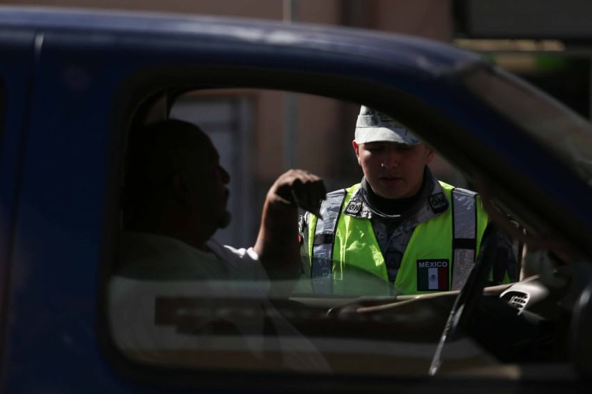 Mexican soldier in uniform and neon emergency personnel vest with a Mexico patch talks to a male driver inside a passenger vehicle as he approaches the border.