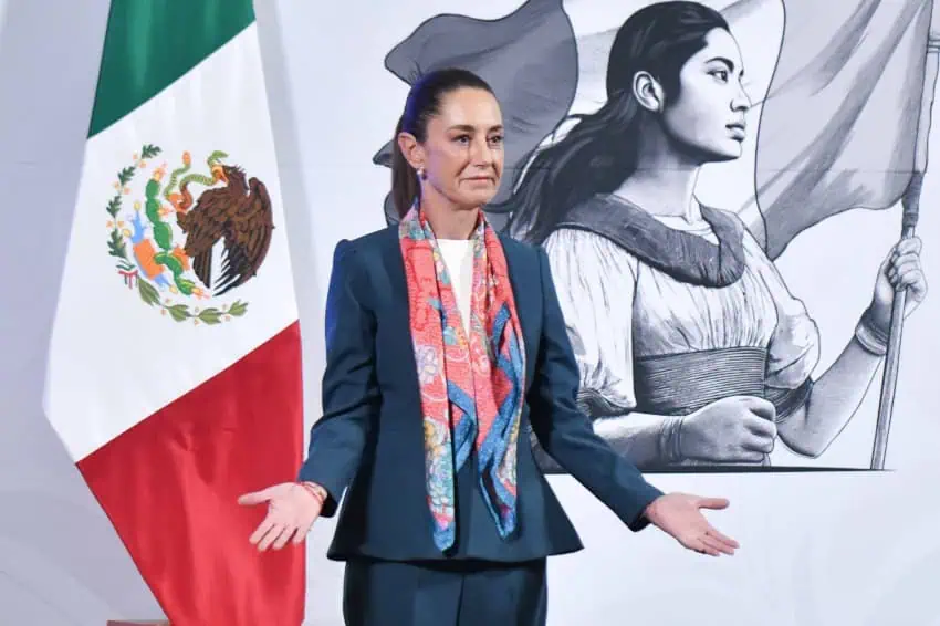 Claudia Sheinbaum at a press conference smiling with her hands outstretched at either side of her body, with her palms upward.