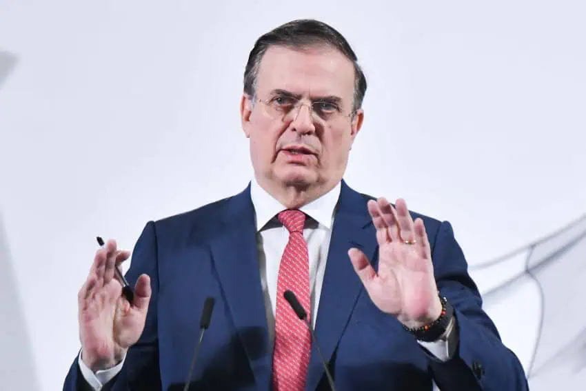 Mexico Economy Minister Marcelo Ebrard at a presidential press conference with his hands in front of his body as he stands behind a podium with microphones. He holds a inexpensive ball point pen in one hand as he speaks.