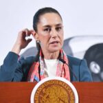 Mexican President Claudia Sheinbaum at the presidential podium touching her right ear with the fingers of her right hand as she speaks to reporters.