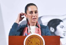 Mexican President Claudia Sheinbaum at the presidential podium touching her right ear with the fingers of her right hand as she speaks to reporters.