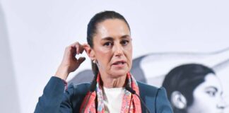 Mexican President Claudia Sheinbaum at the presidential podium touching her right ear with the fingers of her right hand as she speaks to reporters.