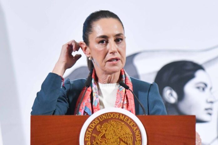 Mexican President Claudia Sheinbaum at the presidential podium touching her right ear with the fingers of her right hand as she speaks to reporters.