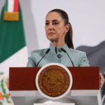 Mexican President Claudia SHeinbaum standing at the presidential podium during her morning press conference. Her expression is grim as she listens to a reporter speaking who is not in the photo.