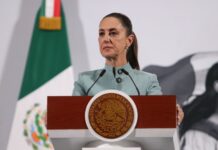 Mexican President Claudia SHeinbaum standing at the presidential podium during her morning press conference. Her expression is grim as she listens to a reporter speaking who is not in the photo.