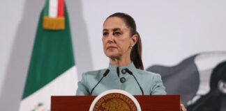 Mexican President Claudia SHeinbaum standing at the presidential podium during her morning press conference. Her expression is grim as she listens to a reporter speaking who is not in the photo.