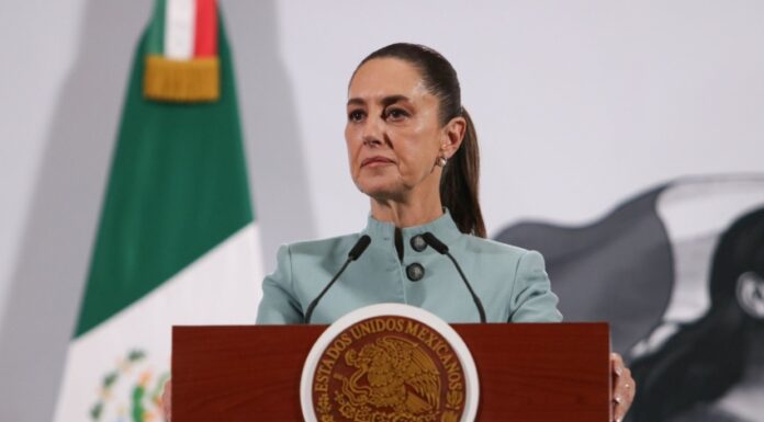 Mexican President Claudia SHeinbaum standing at the presidential podium during her morning press conference. Her expression is grim as she listens to a reporter speaking who is not in the photo.