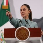 Mexican President Claudia Sheinbaum behind a podium at her daily press conference pointing out at the crowd of reporters as if to pick a person to speak.