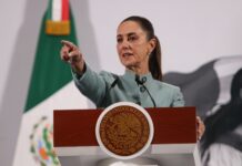 Mexican President Claudia Sheinbaum behind a podium at her daily press conference pointing out at the crowd of reporters as if to pick a person to speak.