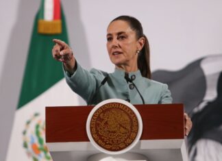 Mexican President Claudia Sheinbaum behind a podium at her daily press conference pointing out at the crowd of reporters as if to pick a person to speak.