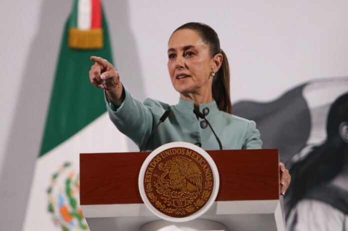 Mexican President Claudia Sheinbaum behind a podium at her daily press conference pointing out at the crowd of reporters as if to pick a person to speak.