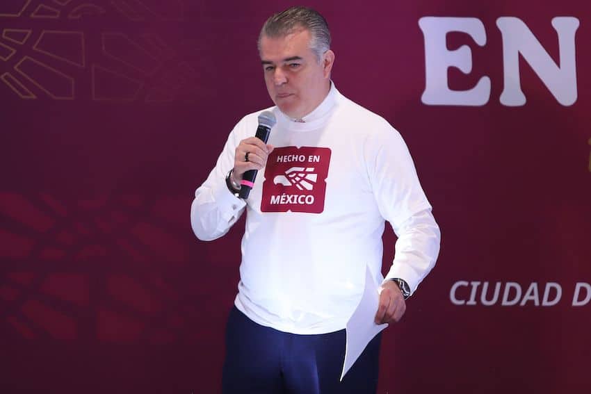 Middle aged man wearing a white sweatshirt with an image of the Hecho en Mexico logo onstage, holding a microphone and with a paper printout in his other hand. He is in mid speech.