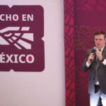 Mexico's Economy Minister Marcelo Ebrard standing on a stage and holding a microphone with the "Hecho en Mexico" seal to the right of him illustrated on a wall.