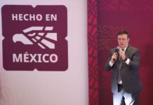 Mexico's Economy Minister Marcelo Ebrard standing on a stage and holding a microphone with the "Hecho en Mexico" seal to the right of him illustrated on a wall.