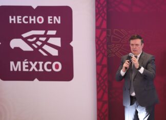 Mexico's Economy Minister Marcelo Ebrard standing on a stage and holding a microphone with the "Hecho en Mexico" seal to the right of him illustrated on a wall.