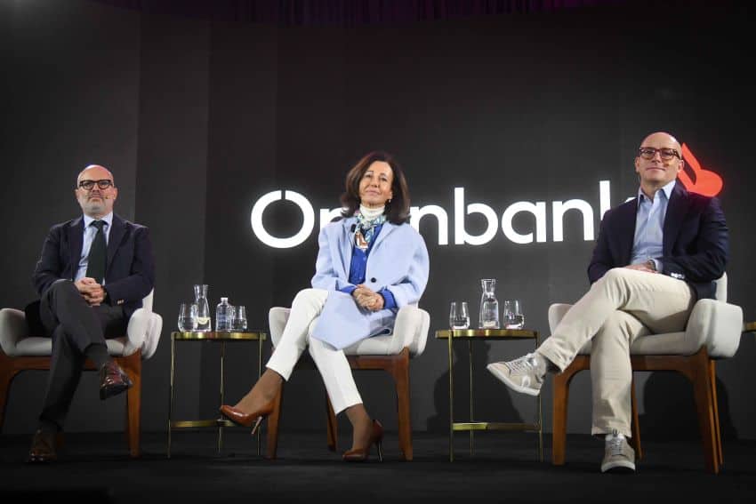 Three people sitting on a stage on upholstered chairs. Between them are gold plated drink tables with bottles of water and glasses. Behind all three is a logo on the wall in white letters for Openbank.