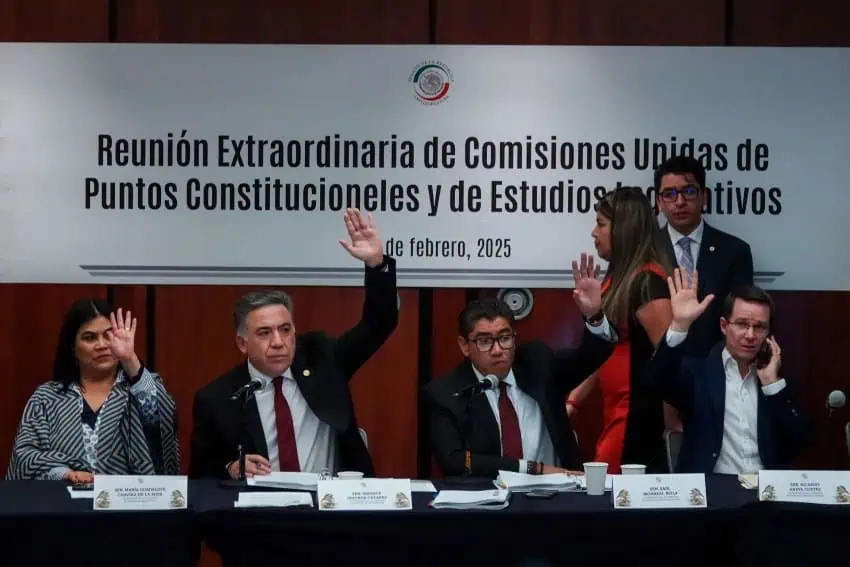 Mexican legislators and officials in the Congress preparing to discuss an anti-nepotism bill. Four people are seated at a banquet style table with their name cards in front of them. They are all raising their hands as two other people from behind them look on or walk by.