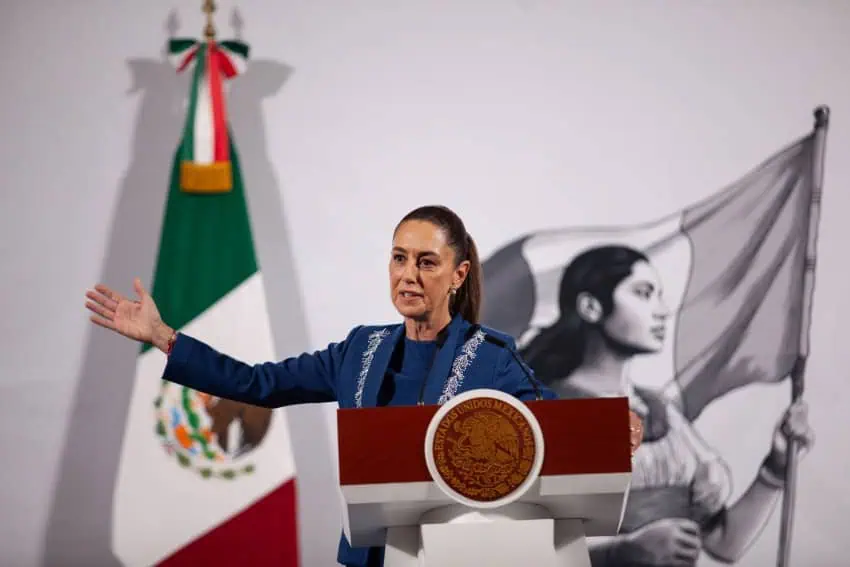 Mexico's President Claudia Sheinbaum at her daily press conference gesturing outward with her right hand while talking to reporters behind a podium at the National Palace.