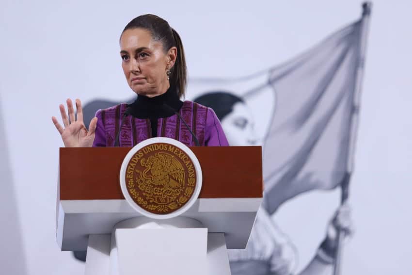 President Claudia Sheinbaum at her daily press conference, standing behind the presidential podium, talking to reporters