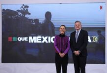 President Sheinbaum at her daily press conference standing and posing for a picture with Netflix CEO Ted Sarandos. They are standing in front of a projection screen with a still of a crew filming a movie