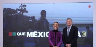 President Sheinbaum at her daily press conference standing and posing for a picture with Netflix CEO Ted Sarandos. They are standing in front of a projection screen with a still of a crew filming a movie