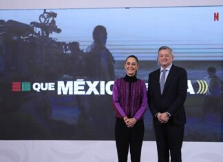 President Sheinbaum at her daily press conference standing and posing for a picture with Netflix CEO Ted Sarandos. They are standing in front of a projection screen with a still of a crew filming a movie