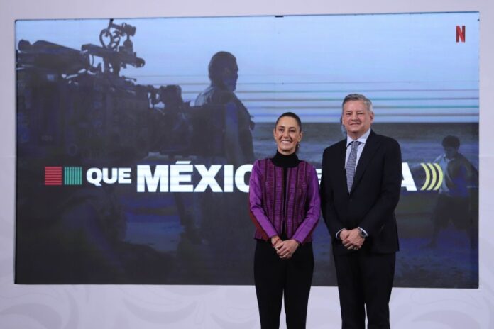 President Sheinbaum at her daily press conference standing and posing for a picture with Netflix CEO Ted Sarandos. They are standing in front of a projection screen with a still of a crew filming a movie