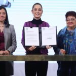 President Claudia Sheinbaum stands at a table in the national palace press briefing room holding open a portfolio with a sheet of paper on each side that contains the text of proposed reforms to the Mexican constitution.