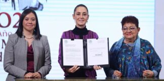 President Claudia Sheinbaum stands at a table in the national palace press briefing room holding open a portfolio with a sheet of paper on each side that contains the text of proposed reforms to the Mexican constitution.