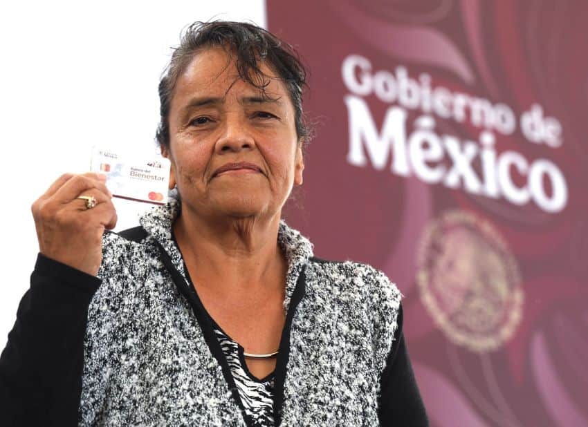 Middle aged Mexican woman holding a government card that says Mujeres Bienestar on it. She is standing on a stage posing for the photo with the card in front of her, with a banner saying "Gobierno de Mexico" in the background.